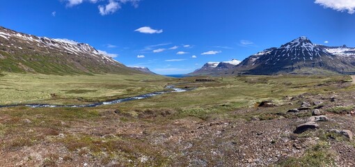panoramic landscape of a mountain river in a barren valley flowing towards a fjord with snow capped mountains in the distance