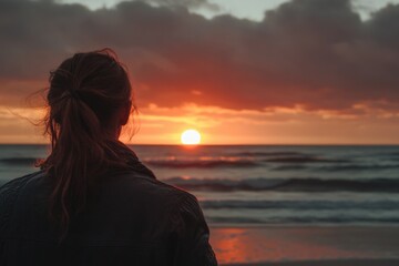 Fototapeta premium A woman stands alone on a beach, gazing out at the setting sun