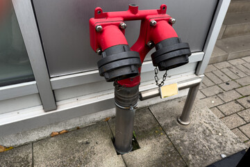 A bright red fire hydrant connection sits on the sidewalk near a building, showcasing two separate...