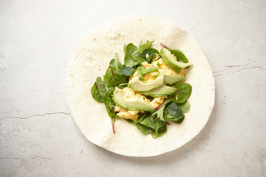 Tortilla wrap on a white surface topped with avocado slices, spinach, and scrambled egg