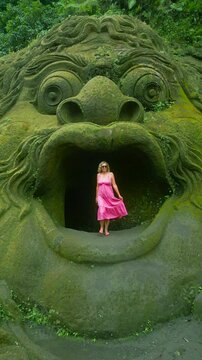 A woman in a pink dress stands inside the moss-covered mouth of a giant stone statue in Ubud on Tegalalang rice terraces.