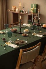 Interior design of warm dining room with table, wreath, christmas tree, green tablecloth, stylish chair, glasses candle with candlestick, brown wall and personal accessories. Home decor. Template.	