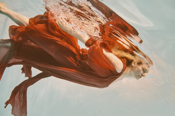 Girl in red dress with long hair swims underwater on light background
