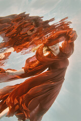 Girl in red dress with long hair swims underwater on light background