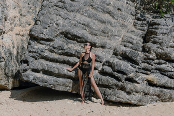 A stylish woman stands confidently by a massive rock formation, showcasing her beach attire while enjoying the warm sun on a beautiful summer day by the sea