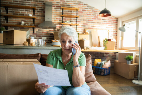 Senior woman making a phone call while reviewing paperwork in living room with moving boxes