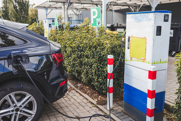 A modern electric vehicle is connected to a charging station in an outdoor public parking area,...