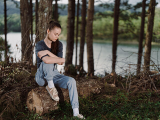 Thoughtful young woman sitting on a log by the lake, wearing casual gray overalls and sneakers, surrounded by trees and a serene natural landscape