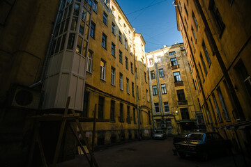 Fototapeta premium A view from the old Saint Petersburg courtyard, looking up towards the sky, framed by worn-out, weathered walls and parked cars below.