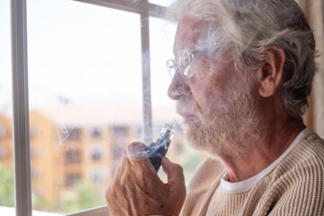 Sad melancholy senior bearded man at home looking out the window smoking electronic cigarette, pensive elderly smoker man using alternative cigarette