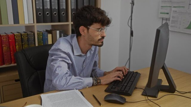 Young man with a beard working indoors on a computer in an office while experiencing neck pain, with books and binders on a shelf in the background