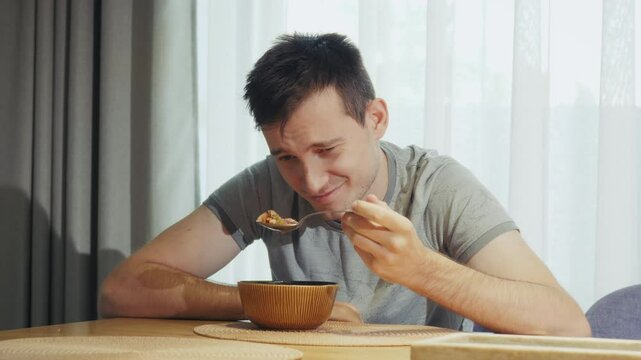 A young man sitting at the dining table, grimacing as he looks at his spoon, struggling to eat a dish he dislikes.