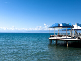A small white pier with blue canopies extends over the blue-green waters of the lake. A bright sunny day, the sea reflects the cloudless sky.