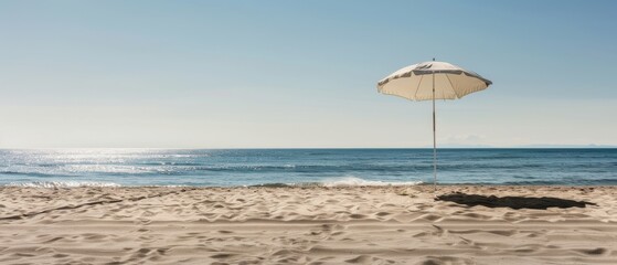 Fototapeta premium A solitary white parasol casts a long shadow on the golden sandy beach, with the tranquil blue sea stretching to a clear horizon under the bright sky.