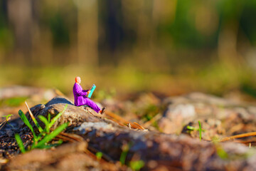 Miniature Figurine in Purple Suit Reading on a Mossy Log in the Woods