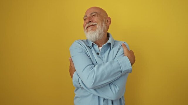 Man, senior bald gentleman wearing denim shirt standing and hugging himself. a happy, confident, smiling, and positive expression, embracing self love over an isolated yellow background