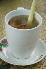 A cup of hot ginger tea (Zingiber officinale) with sugar and lemongrass (Cymbopogon citratus), served in a white cup and saucer. A popular Indonesian drink. Blurred woven mat background.