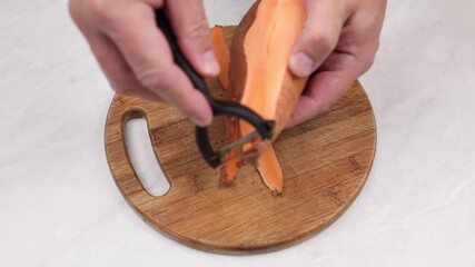 Close up footage showcasing a sweet potato being peeledby man ahnds with thin slices scattered on a wooden cutting board, highlighting the vibrant orange color and texture of the vegetable