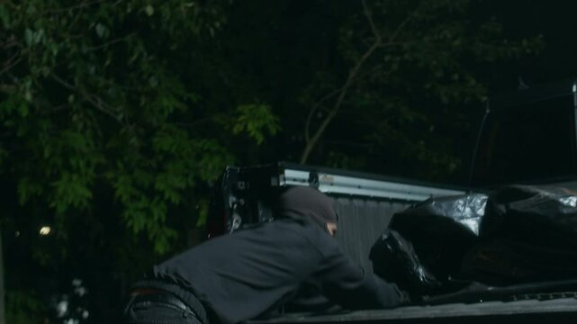 Two unrecognizable offenders in dark clothes and masks pulling corpse in black body bag along ground then throwing it at truck bed, getting rid of evidence after committing homicide at night
