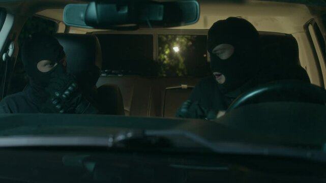 Two dangerous robbers putting on black masks on their heads and face and getting out of car while committing crime at night