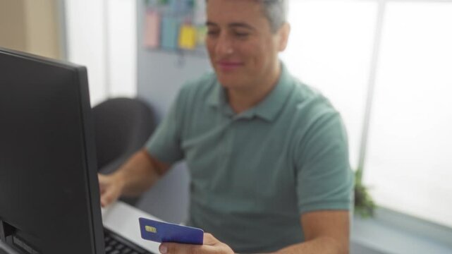 Man in green shirt holding credit card while working at desk in modern office, typing on keyboard with computer monitor, smiling confidently indicating secure online shopping or banking