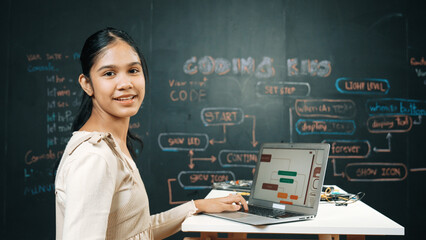 Caucasian girl working on laptop while smiling to camera at blackboard with engineering code and...