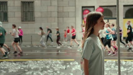 Teenager standing near building, observing marathon runners on wet street with scattered plastic cups, highlighting environmental impact of sports events