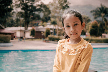 A young girl poses near an outdoor swimming pool with a calm expression, wearing a yellow blouse. Trees and mountains are visible in the background, capturing a serene and sunny day.