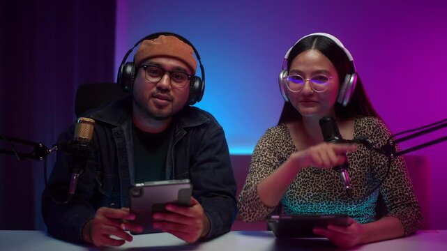 Two young co host creator radio being live streaming using microphones and wearing headphone at home studio broadcast room, cheerful vloggers broadcast recording podcast and speaking to audiences.
