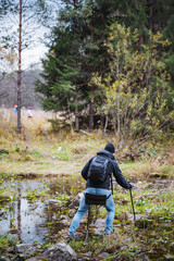 Obraz premium A man, who is carrying a backpack, is carefully crossing a stream that runs through the beautiful woods filled with trees and grass, surrounded by a peaceful natural landscape