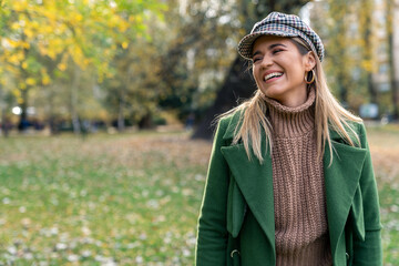 Smiling woman in trendy green coat and checkered hat enjoys a leisurely stroll, the golden autumn...