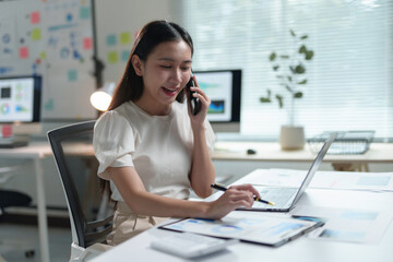 Fototapeta premium Smiling businesswoman working with documents and laptop while talking on the phone in modern office