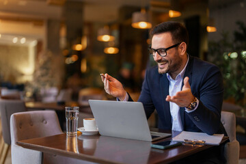 A mid adult Caucasian man in sharp business attire conducts a video call on his laptop, gesturing actively while surrounded by the modern decor of a bustling cafe.