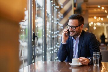 A cheerful mid-adult Caucasian man in professional attire talks on his smartphone while sitting with a coffee cup at a well-lit cafe table.