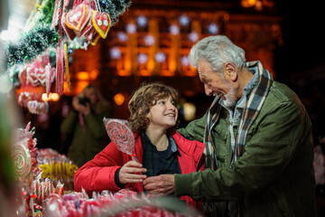 Elderly man with a joyful boy selecting traditional hard candy at a decorated market stall,...