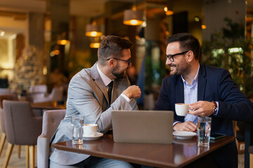 Two mid adult Caucasian men dressed in business attire engage in a discussion with a laptop and coffee mugs on their table, inside a well-lit modern cafe.