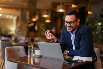 A mid-adult Caucasian male in a smart suit engages in a video call on his laptop while seated at a...
