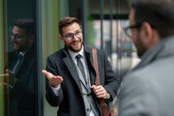 Two professional mid adult men in business attire engage in a constructive conversation outside a modern building, exchanging ideas and strategies.