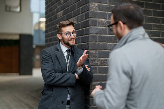 Two mid adult Caucasian men in smart business attire engage in a conversation against an urban backdrop, demonstrating a collaborative professional exchange in an outdoor setting.
