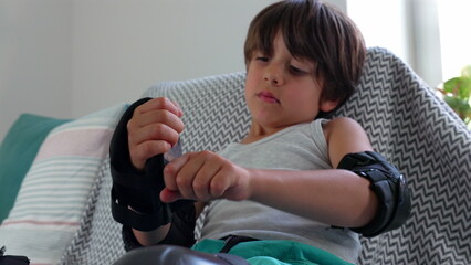 Young boy adjusting his elbow pad while sitting on the couch, focusing on making sure it fits correctly. concentration and patience in getting ready for an active outdoor adventure