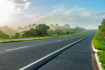 Paved dual-lane highway in perspective, with no cars, early in the morning, with a bit of fog and...