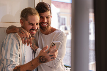 Happy gay couple laughing and using smartphone at home