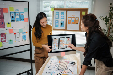Two web designers discussing user interface and user experience design for a mobile application using a desktop computer and sticky notes