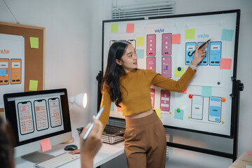Asian women working together as web designers in an office, pointing at whiteboard with uxui mobile app wireframe