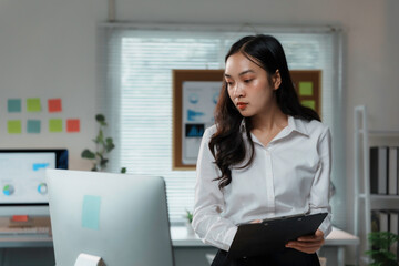 Focused asian businesswoman holding clipboard and reviewing information on computer screen in modern office