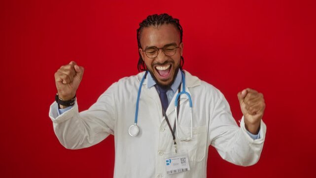 Young man celebrating success over a vibrant red background wearing a white coat with a stethoscope around his neck and clenching his fists with joy and excitement.