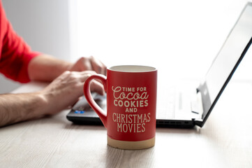 Red mug with festive text next to a person working on a laptop, capturing a cozy, holiday-themed workspace atmosphere