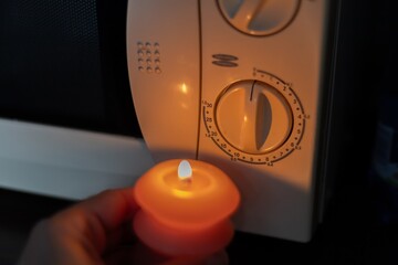 Burning candle in hand near microwave oven in dark. Power outage due to war in Ukraine