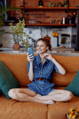Relaxed woman sitting on orange couch with phone in hand and plant in front of her