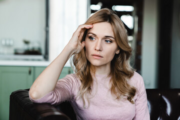 Close up shot of Caucasian unhappy worried woman sitting on couch, touching forehead, feeling...